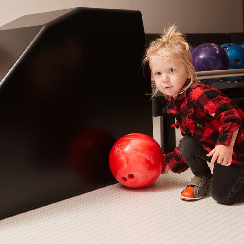 Toddler in plaid shirt holding red bowling ball at bowling alley